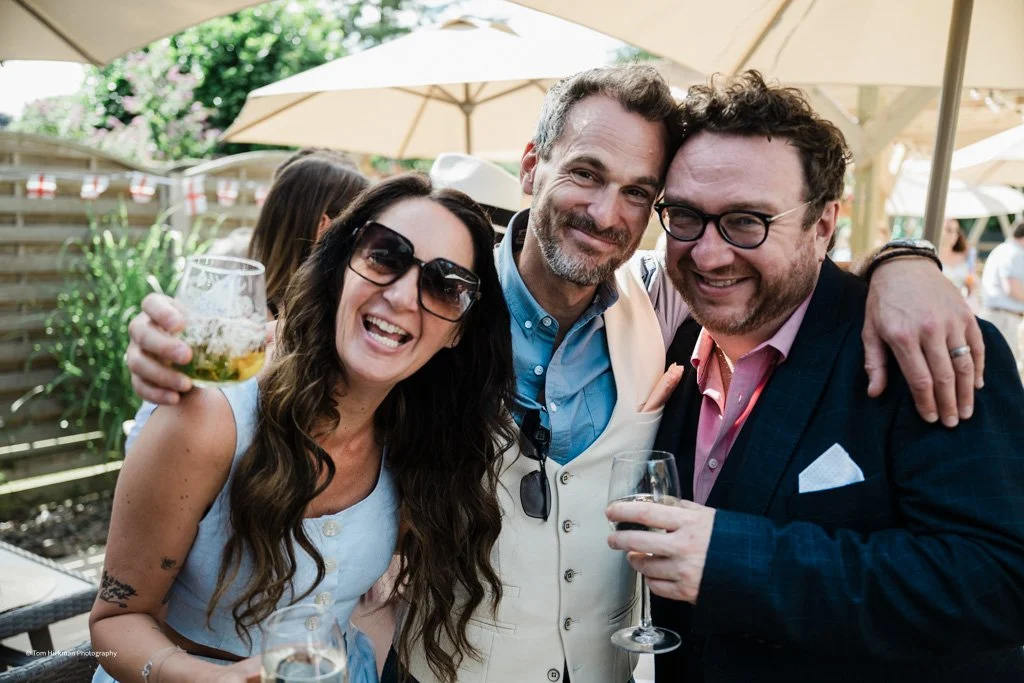 Three people smiling and celebrating outdoors, holding drinks, with umbrellas and a wooden fence in the background.