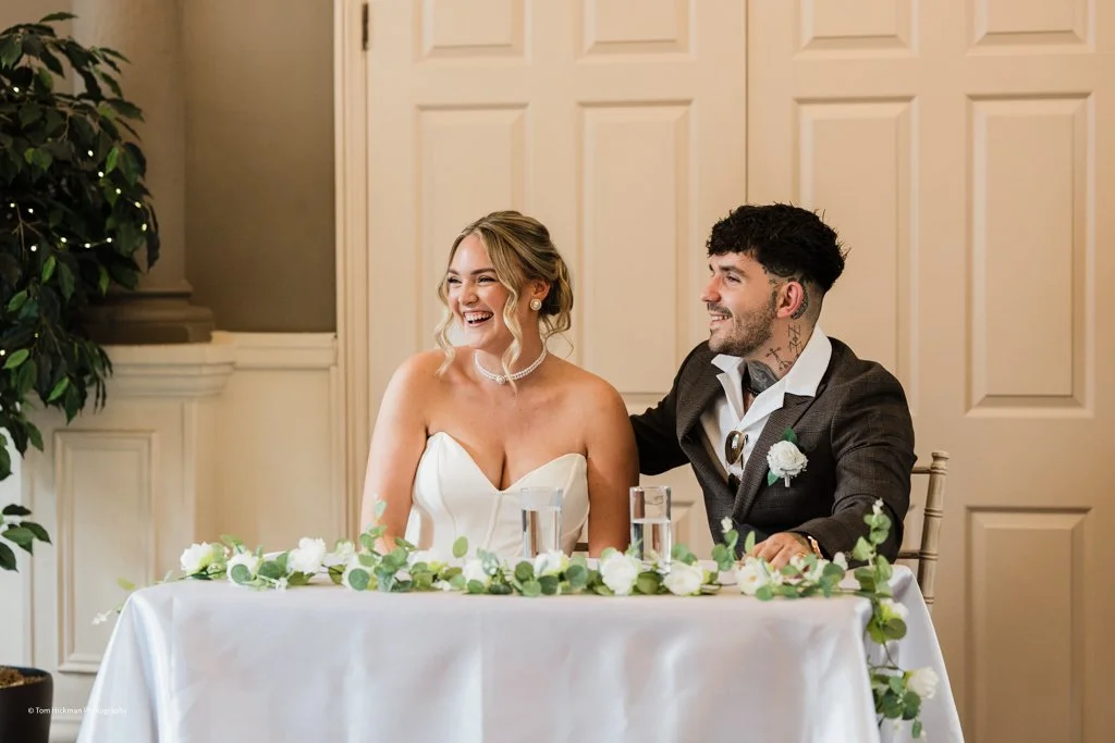A bride and groom sitting at a wedding reception table, smiling and laughing. The bride is wearing a strapless white wedding dress and the groom is wearing a dark suit with a white shirt and a boutonniere.