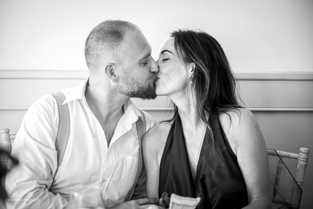 A man and a woman sharing a kiss, sitting close with their eyes closed, in an indoor setting.