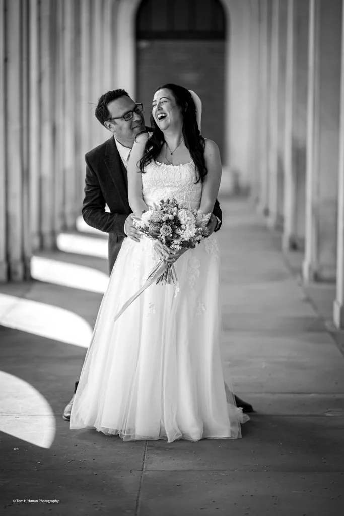 A joyful bride holding a bouquet, laughing with a groom standing behind her, in an elegant indoor setting with curtains.