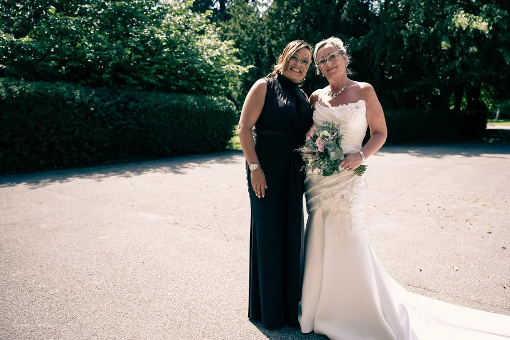Two women smiling and standing outdoors on a sunny day, one in a wedding dress holding a bouquet, and the other in a dark formal dress.