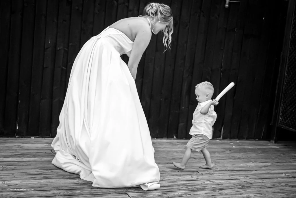 A woman in a long white dress bending down to a young boy holding a baseball bat, outdoors on a wooden deck, with a dark wooden fence in the background.