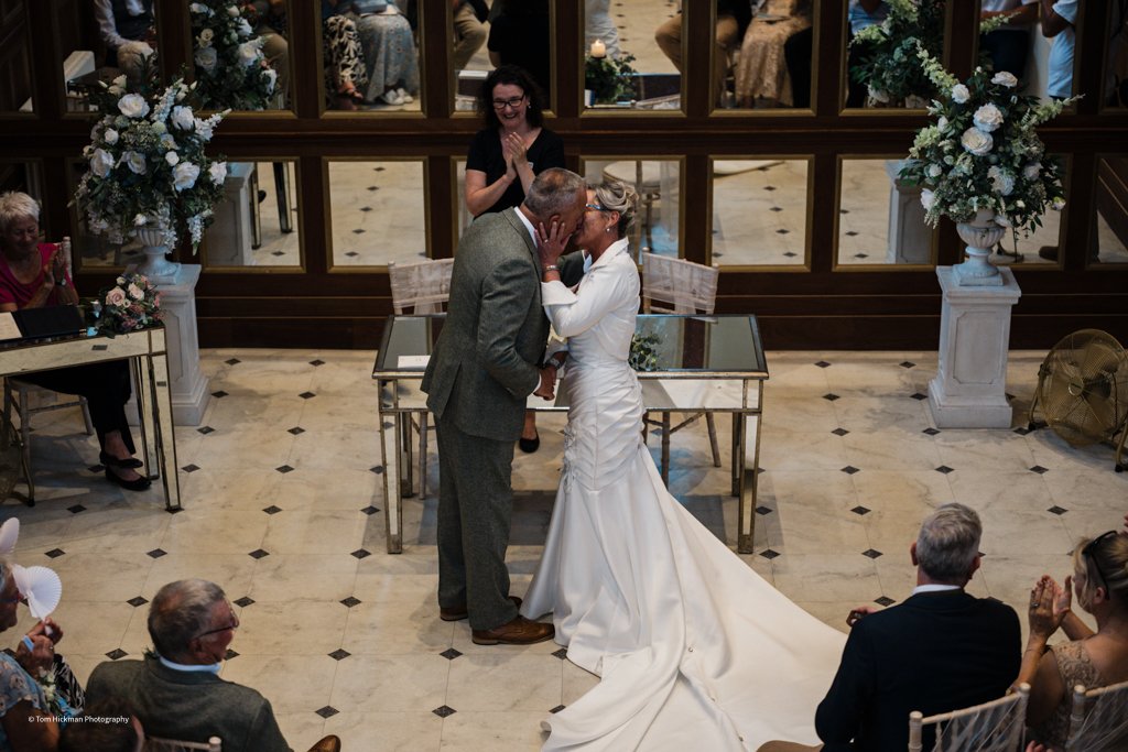 A bride and groom share a kiss during their wedding ceremony in a decorated indoor venue, with guests seated and smiling around them.