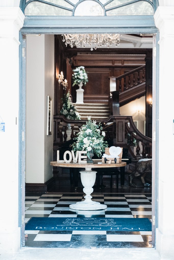 Decorative love-themed display in a hotel lobby with a floral arrangement, wooden table, and large letters spelling 'LOVE', at the Bougainvillea Hotel.