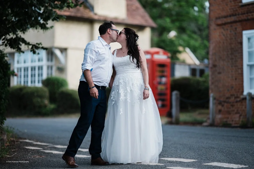A couple in wedding attire sharing a kiss outdoors in a village street setting.
