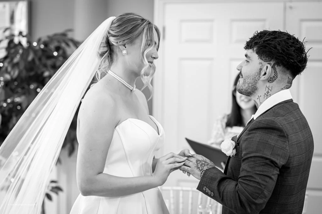 A bride and groom exchanging rings during their wedding ceremony indoors, with a woman smiling in the background.