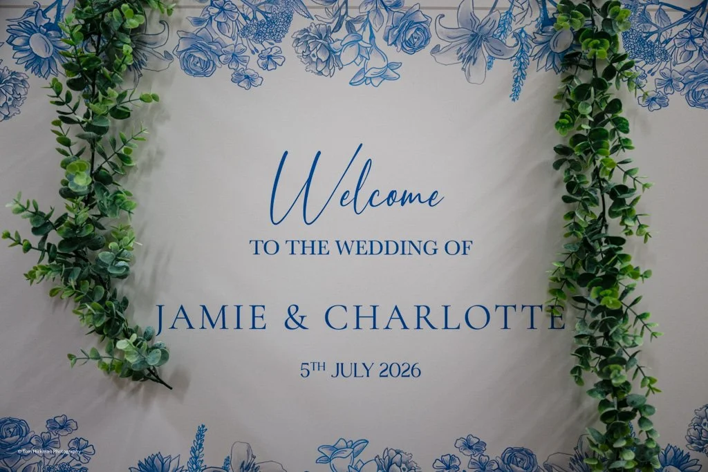 Wedding welcome sign with blue floral border, featuring green leafy garlands framing the sign, which reads: "Welcome to the wedding of Jamie & Charlotte, 5th July 2026."