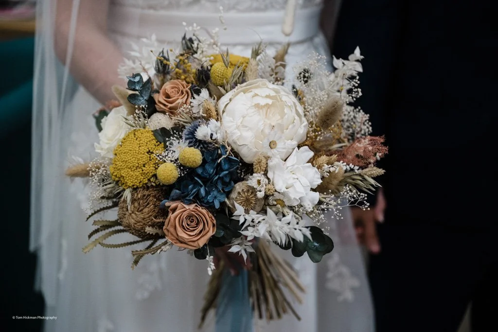 A bridal bouquet with white peonies, blush roses, yellow billy balls, dried grasses, and greenery, held by a bride in a white dress.
