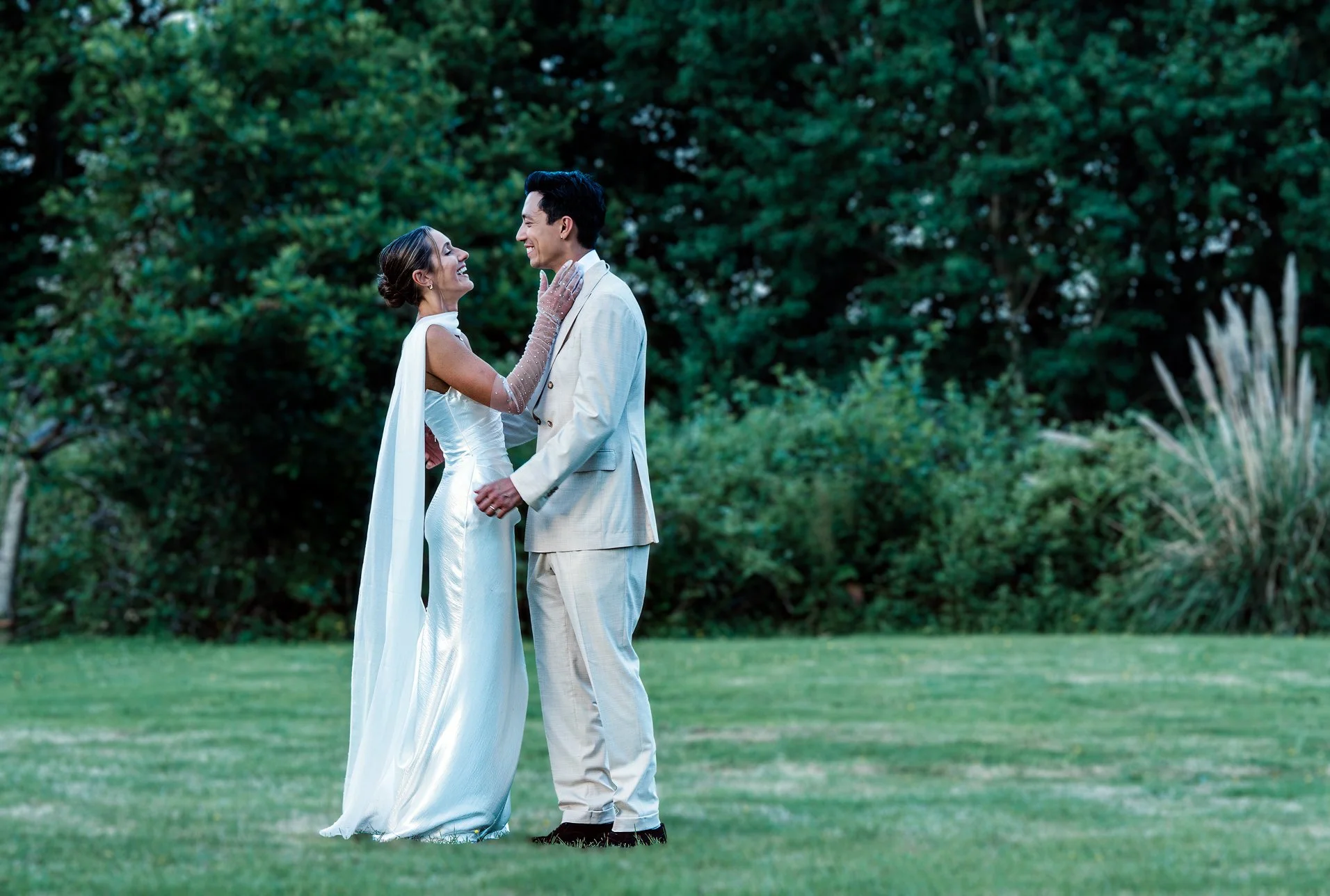 A bride and groom in wedding attire sharing a joyful moment outdoors on a grassy lawn with trees in the background.