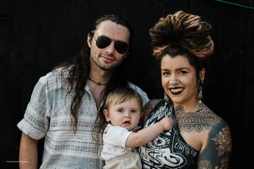 A family of three, a man, a woman with tattoos and a child, standing close together against a dark background.
