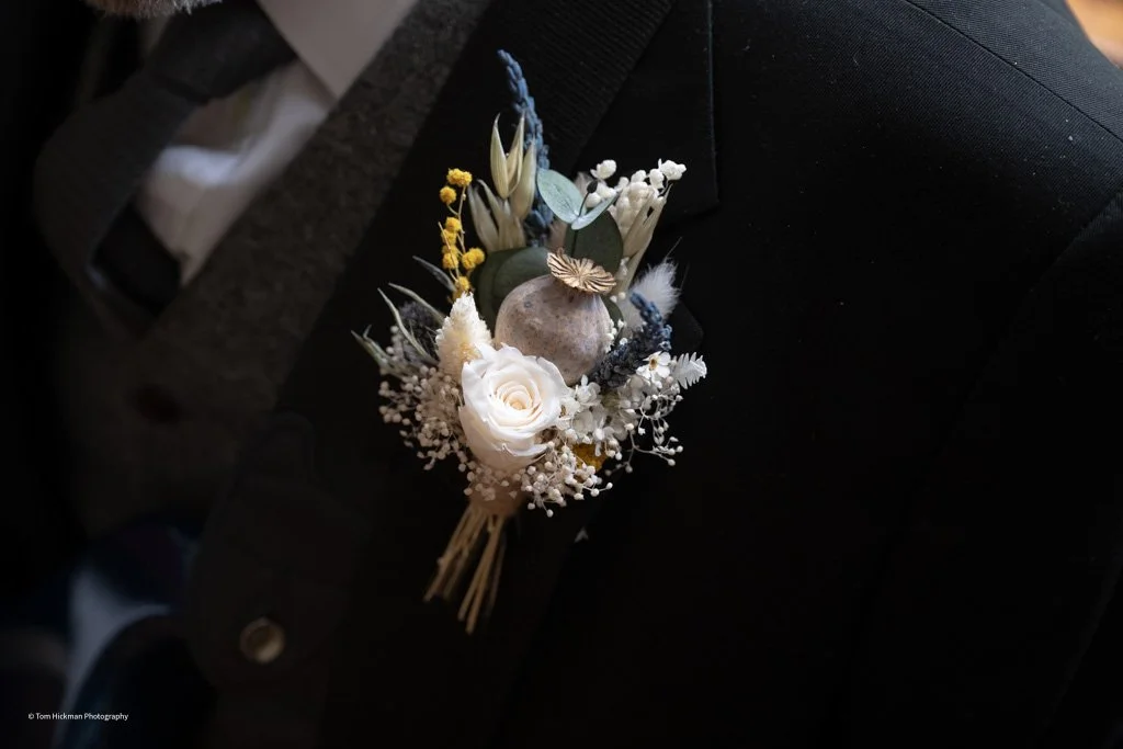 A boutonniere with white roses, dried flowers, greenery, and small decorative elements pinned to a man's dark suit jacket.