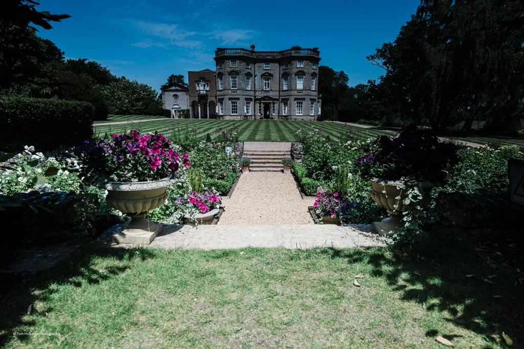 A grand mansion with a panoramic lawn, a garden filled with colorful flowers, and a gravel pathway leading up to the mansion's entrance.