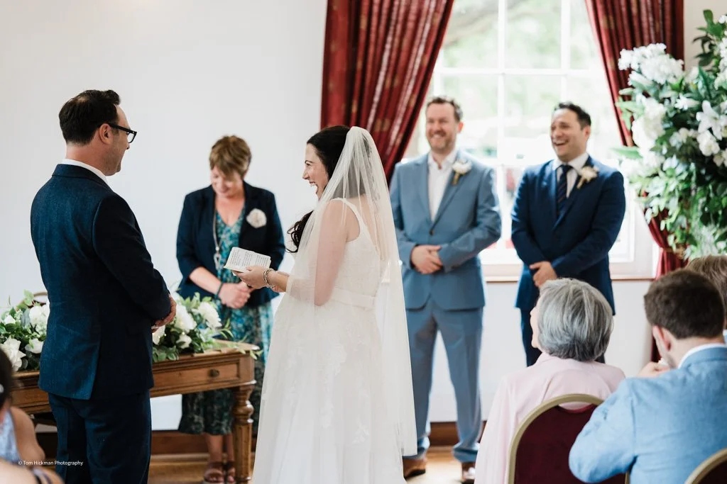 Bride and groom exchanging vows during wedding ceremony indoors with guests and officiant, decorated with flowers and red curtains.