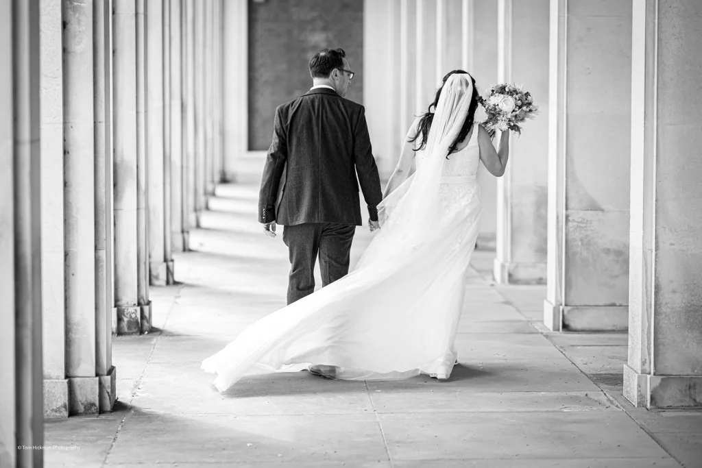 Black and white photo of a newlywed couple walking hand in hand through a columned walkway. The bride is wearing a long wedding gown with a veil and holding a bouquet, while the groom is in a dark suit with glasses.
