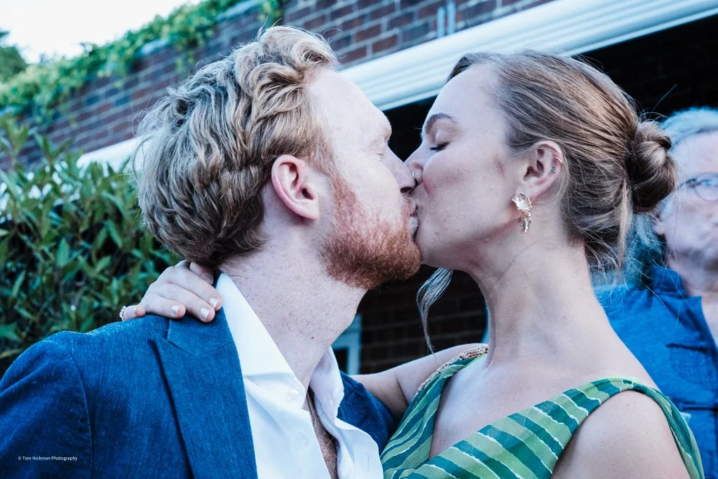 A couple kissing outdoors, with a man in a blue suit and a woman in a striped dress, in front of a brick wall and green plants.