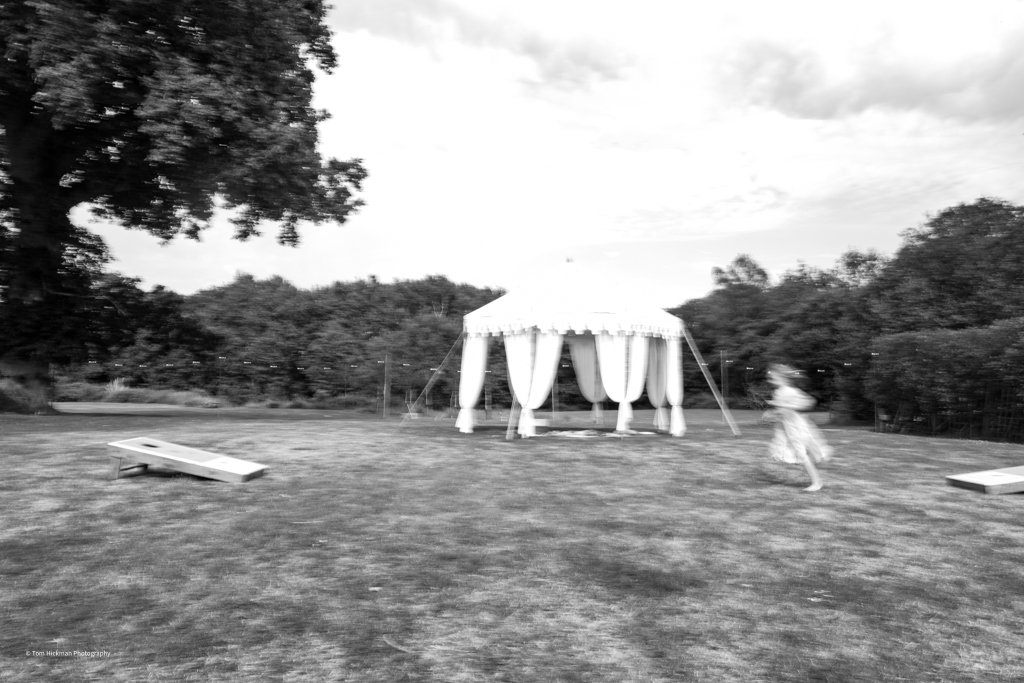 A large white tent with draped fabric in an open grassy area, with a young girl running near it and two cornhole game boards on the ground, surrounded by trees.