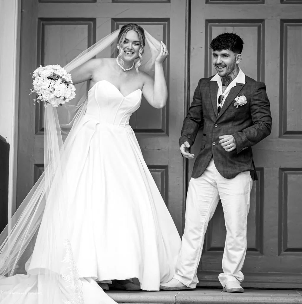 A bride and groom smiling and laughing during their wedding photos in front of a wooden door. The bride wears a strapless white wedding gown and veil, holding a bouquet of flowers. The groom is dressed in a checked blazer, light-colored trousers, and