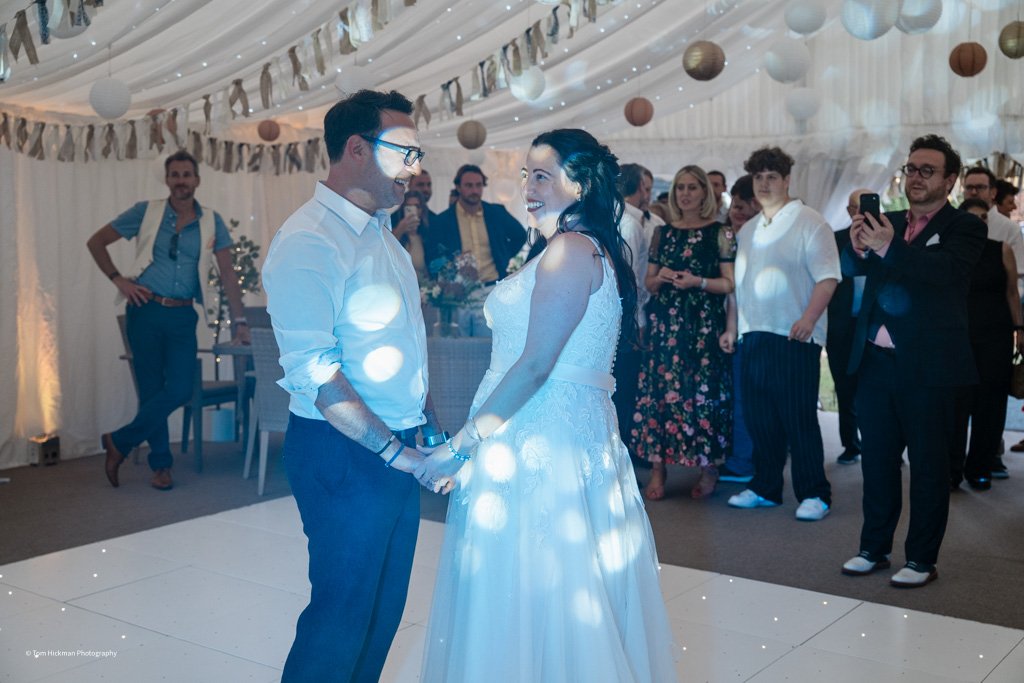 A bride and groom holding hands and smiling at each other during their wedding reception in a decorated tent with hanging lights and paper lanterns, surrounded by wedding guests.