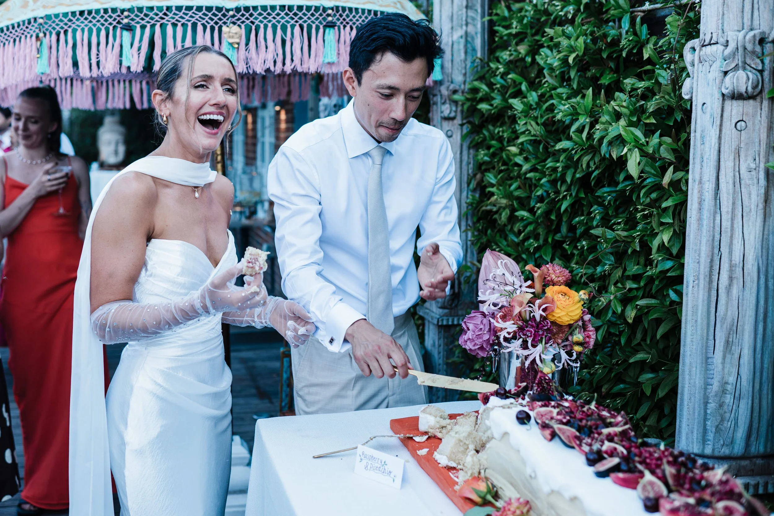 A bride and groom at their wedding reception cutting a cake decorated with flowers, with guests and greenery in the background.
