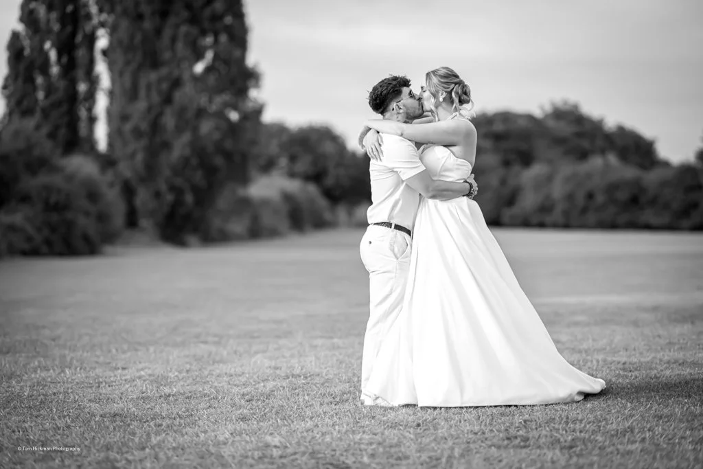 A couple on their wedding day are embracing and about to kiss on a grassy field with trees in the background.