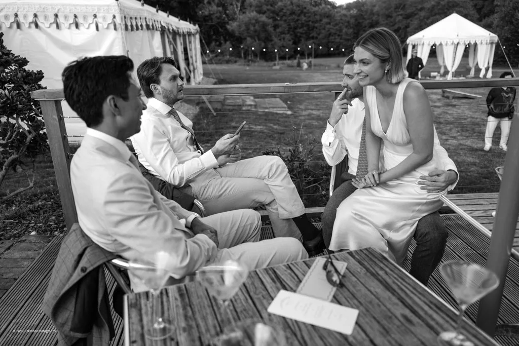 Four people dressed in formal attire sitting outdoors at a wedding reception, with two men and one woman sitting on a bench and a woman sitting on a man's lap, chatting and smiling. There are tents and a grassy area in the background.