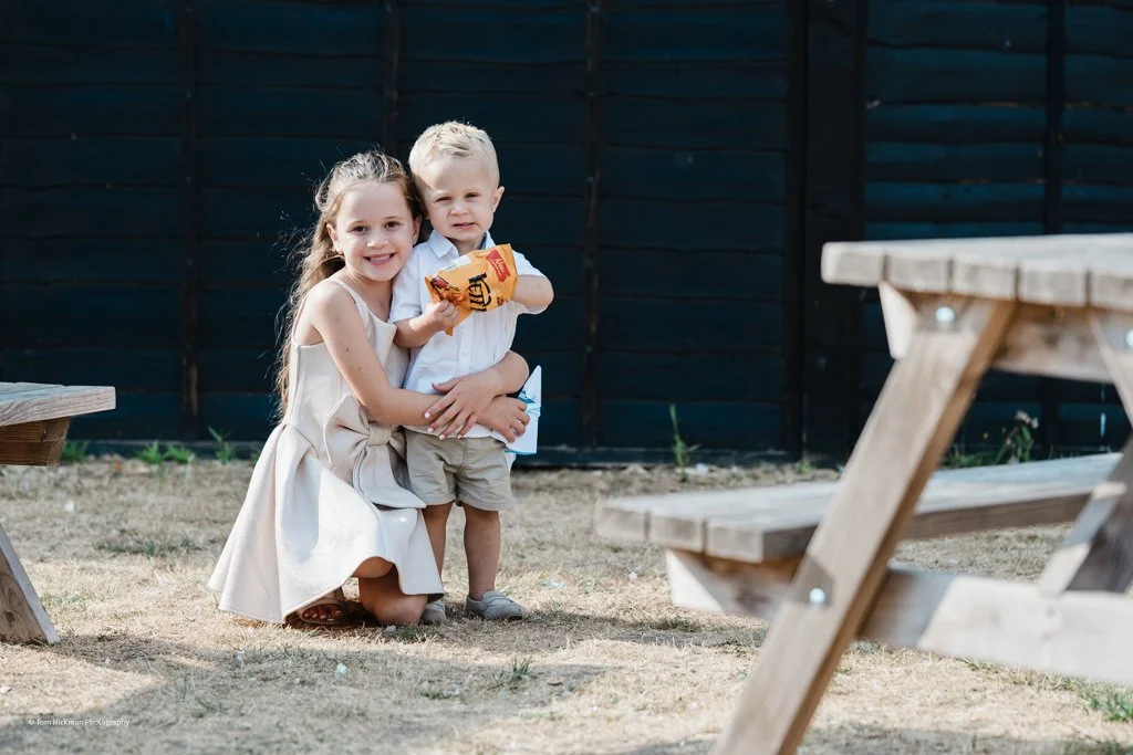 A young girl and boy hugging outdoors near wooden picnic tables with a dark wooden fence in the background.