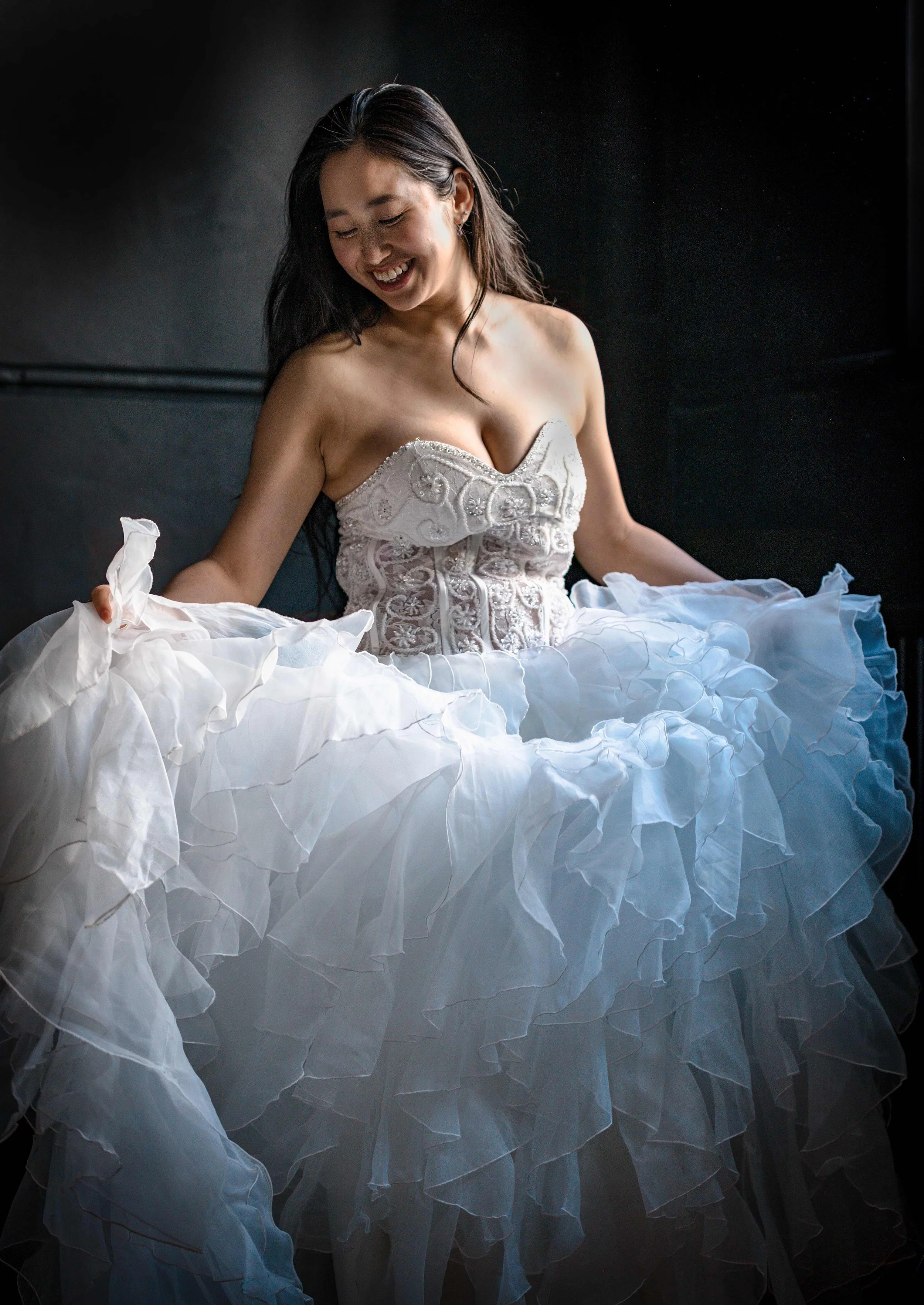 A woman with dark hair smiling and looking down, wearing a white strapless dress with intricate embroidery, sitting among white ruffled fabric pieces against a dark background.