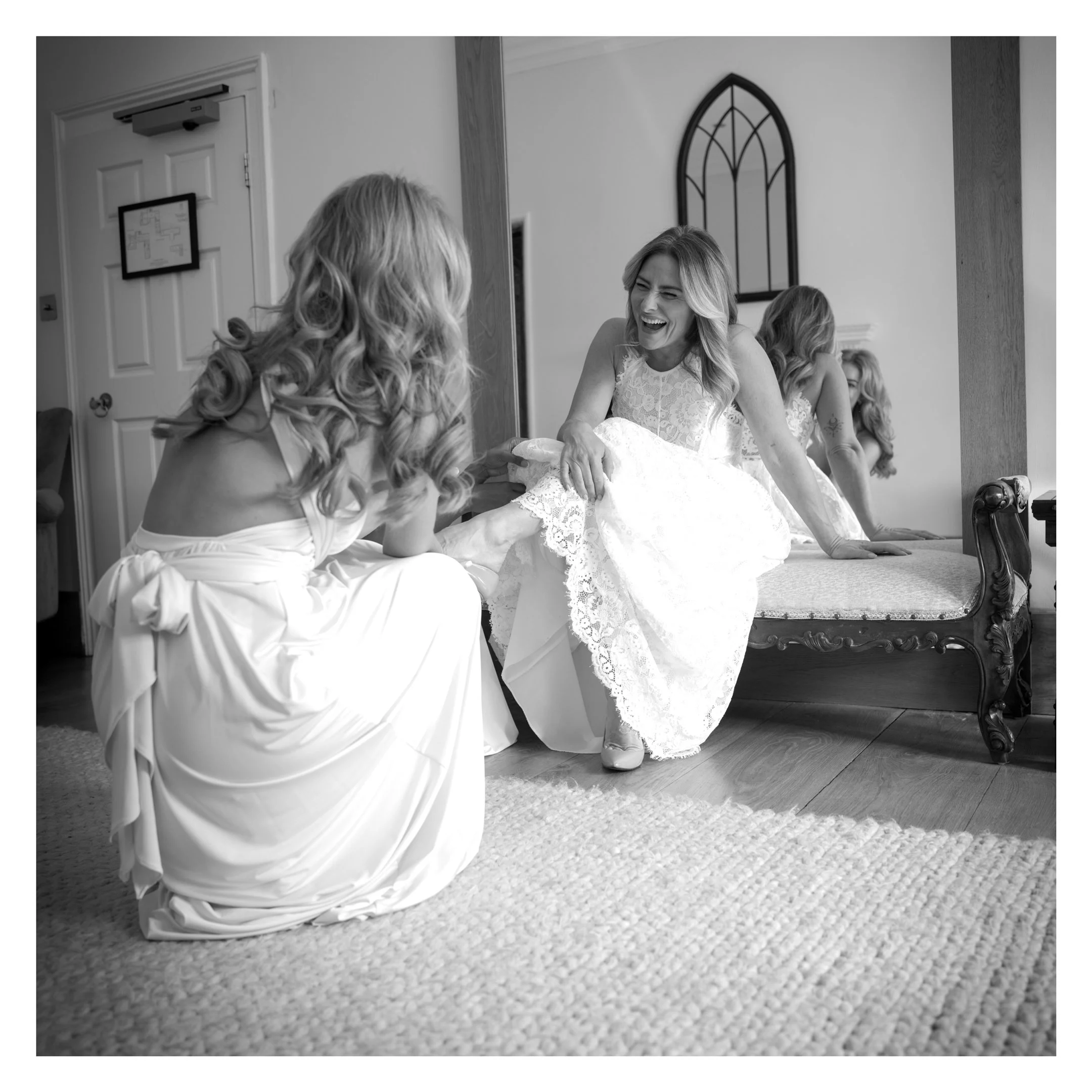 A woman in a lace dress laughing and leaning over a table with three other women, in a room with a mirror, a door, and a framed map on the wall.