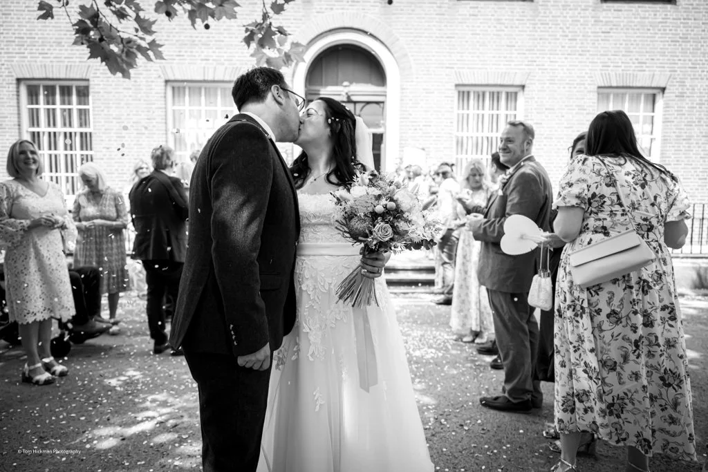 A couple is kissing during their wedding outside a building, with guests in the background.