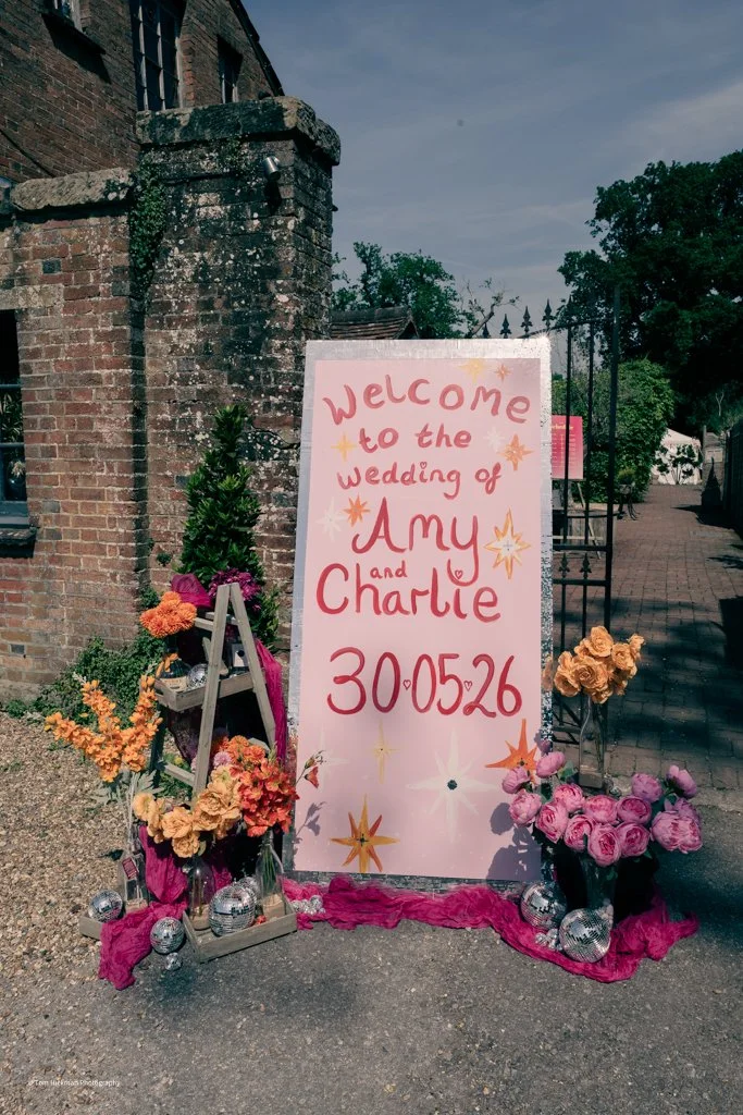A welcoming sign for the wedding of Amy and Charlie, with the date 30.05.26, surrounded by colorful flowers and decorations outside a brick building.