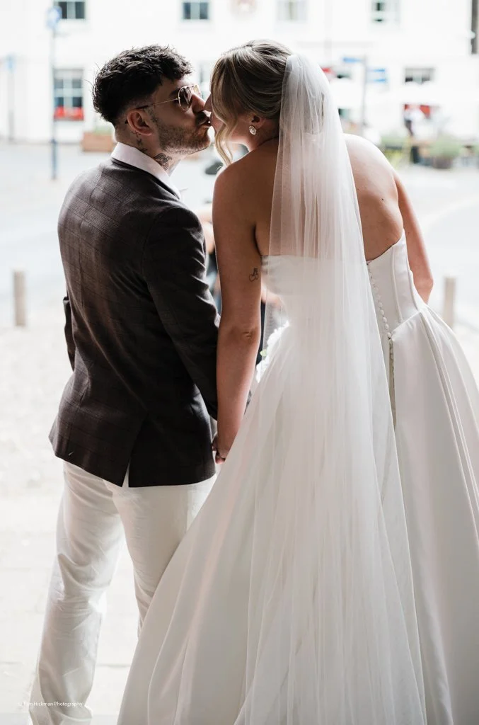 A bride and groom share a kiss outdoors during their wedding, with the bride in a white strapless gown and veil, and the groom in a dark blazer and light pants.