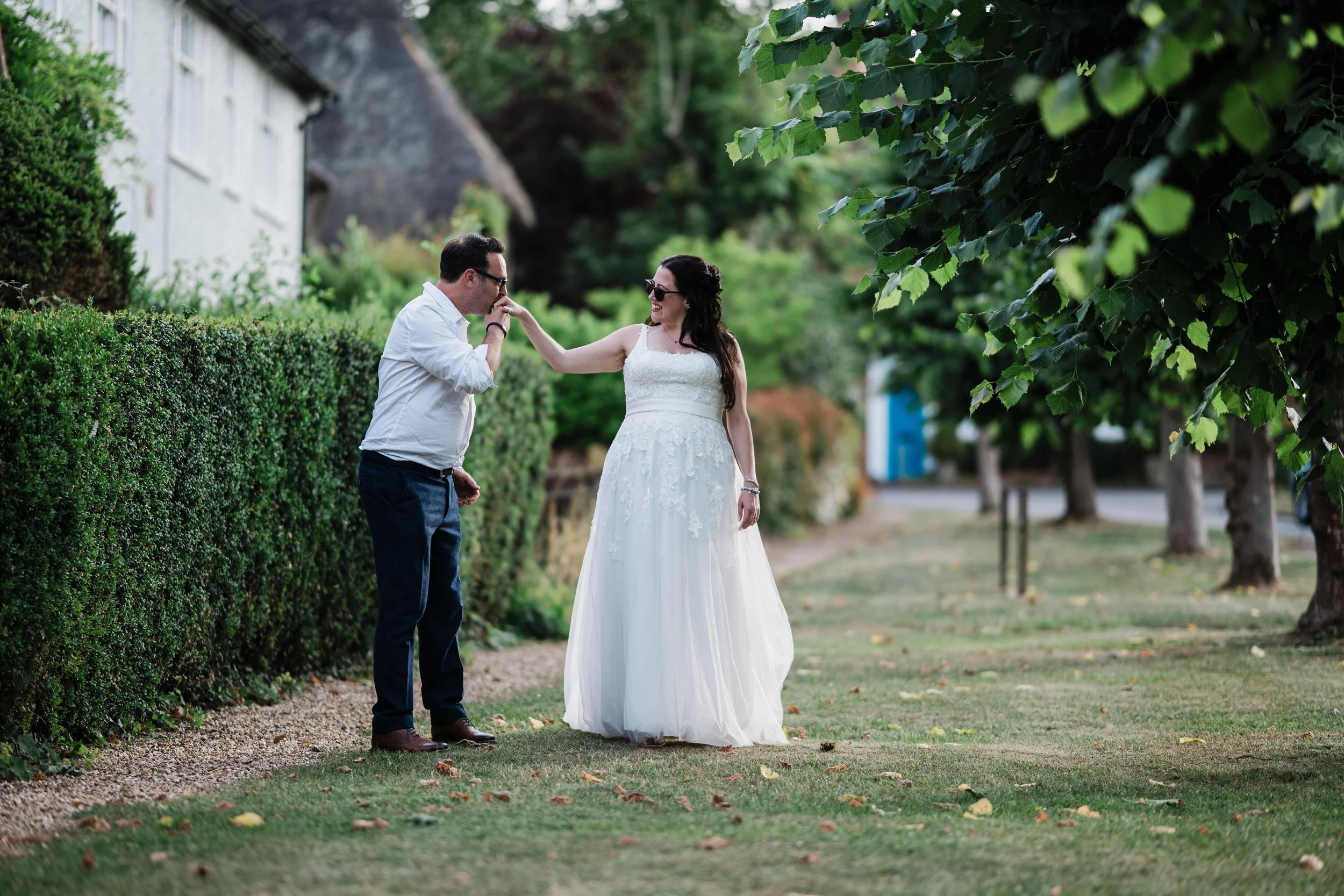 A bride and groom stand outdoors on a grassy area, with the groom kissing the bride's hand. The bride is wearing a white wedding dress and sunglasses, while the groom is dressed in a white shirt and dark pants. They are surrounded by trees and a hedg
