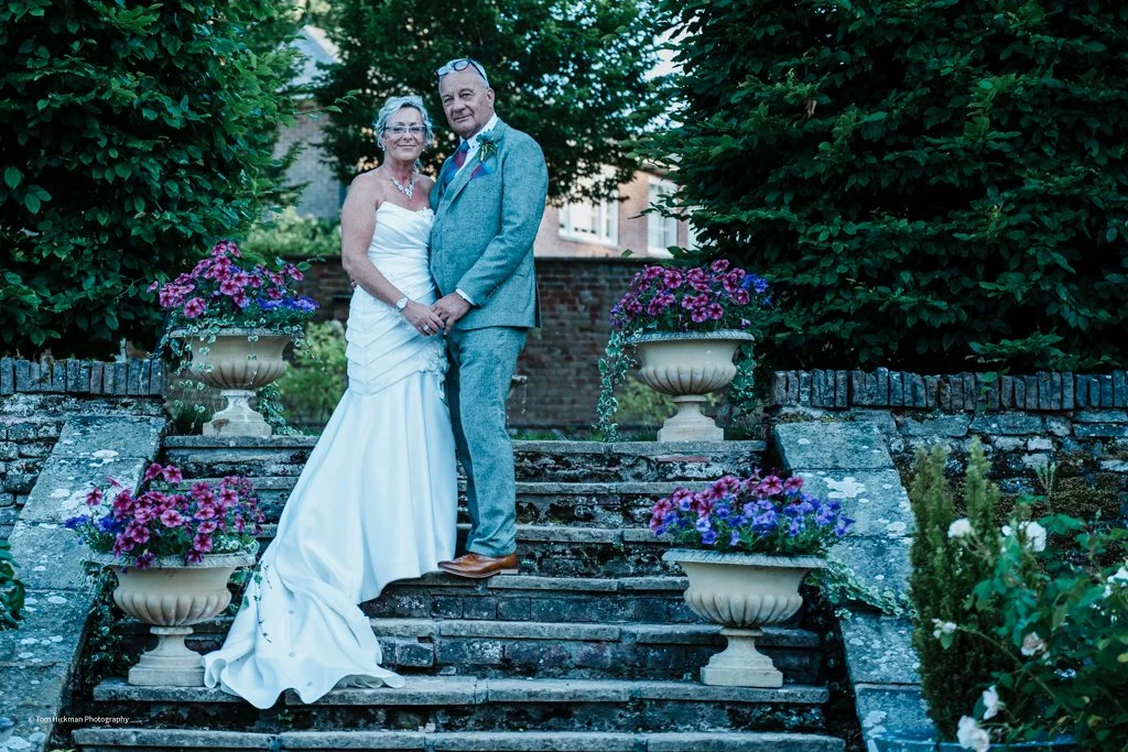 A bride and groom standing on stone steps in a garden, surrounded by large planters with pink and purple flowers, green bushes, and trees in the background.