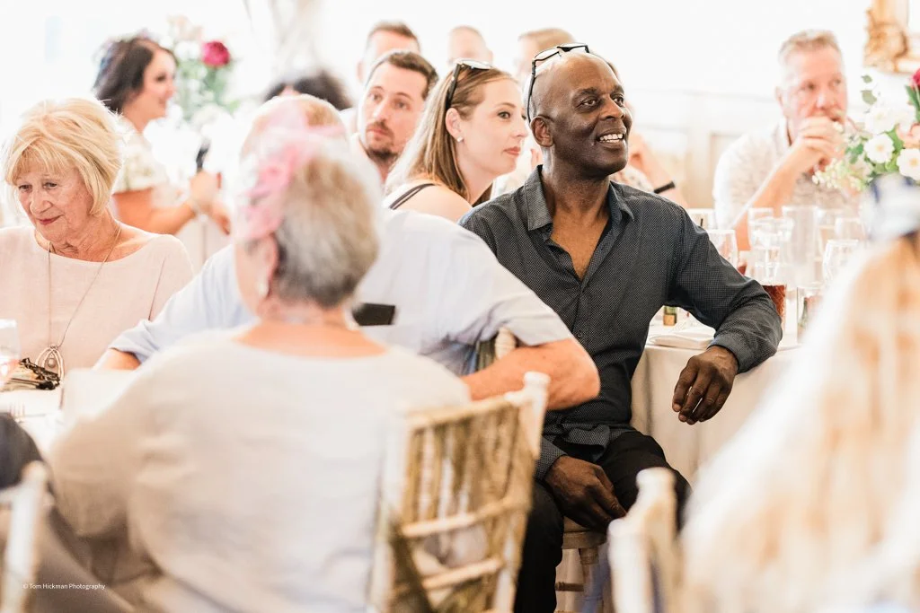 Group of people sitting at a banquet table during a wedding or celebration, listening attentively.