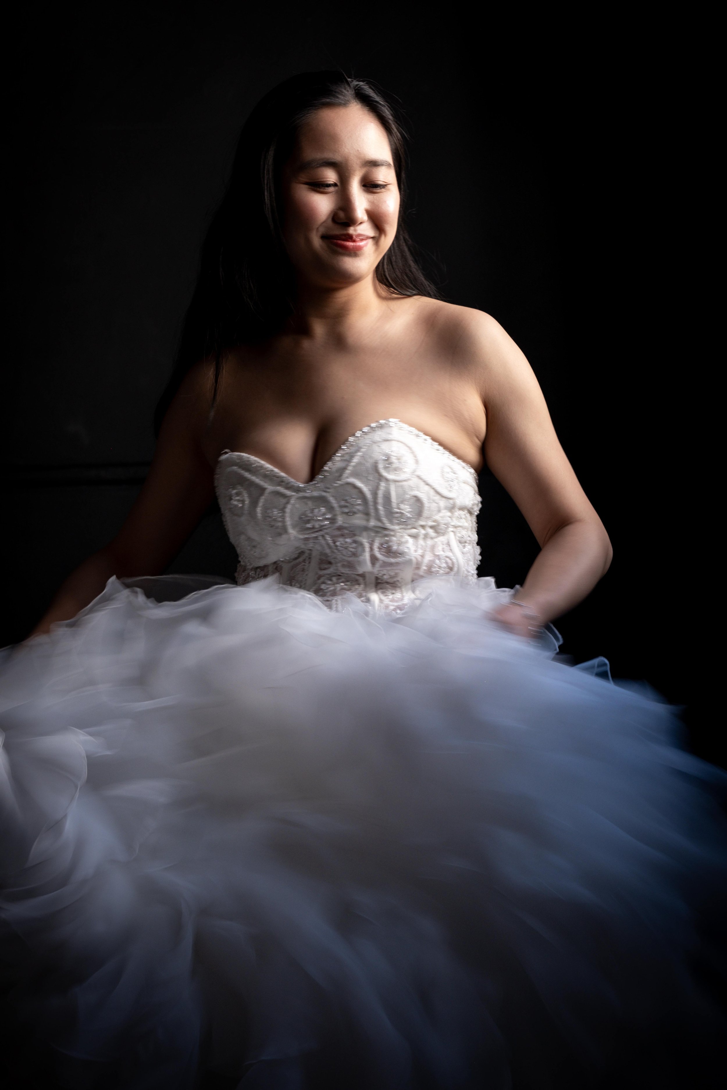 A woman in a white strapless dress with intricate embroidery and tulle skirt, smiling with closed eyes against a dark background.