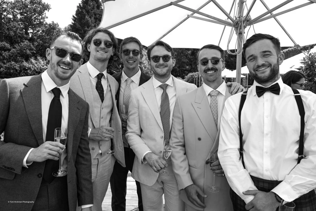 Group of seven men dressed in suits and tuxedos, wearing sunglasses, at an outdoor event under a large umbrella, smiling and holding champagne glasses.