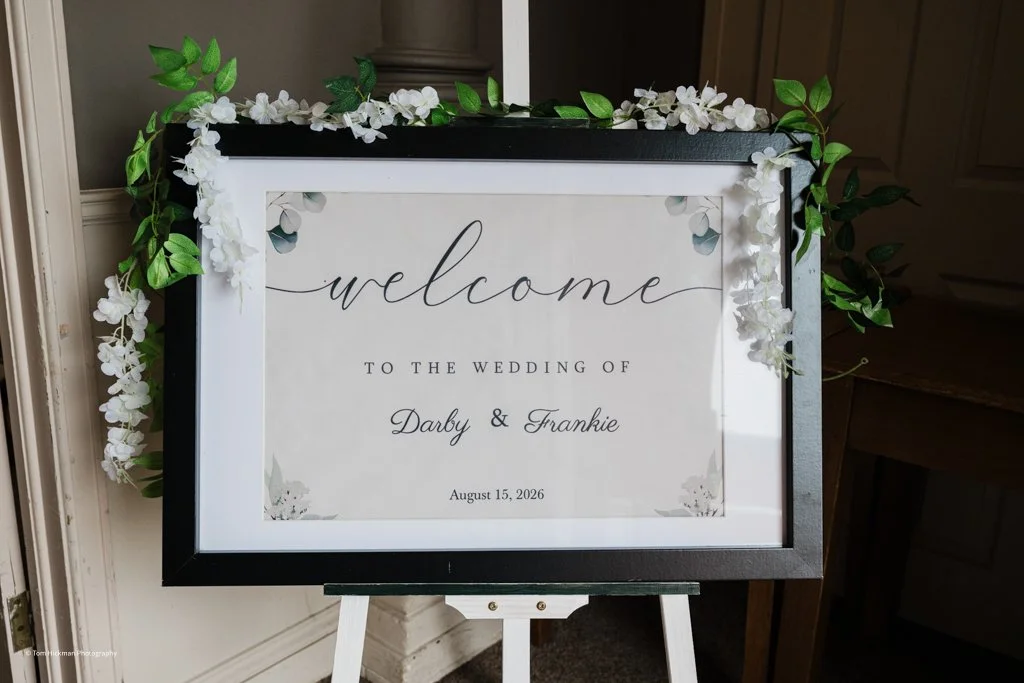 Wedding welcome sign decorated with white flowers and green leaves, featuring the names Darby and Frankie, and the date August 15, 2026.