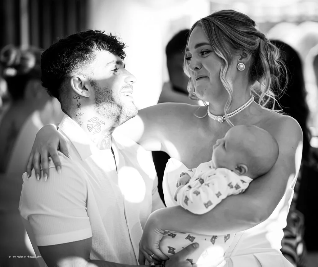 A black and white photo of a man and woman smiling at each other while holding a baby. The woman is wearing jewelry, and the man has tattoos on his neck. The woman is gently holding the baby, who is looking up. They are indoors, and the background sh