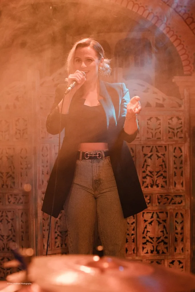 Woman with brown hair, wearing a black jacket and high-waisted jeans, speaking or singing into a microphone on stage with a decorative wood background and warm lighting.