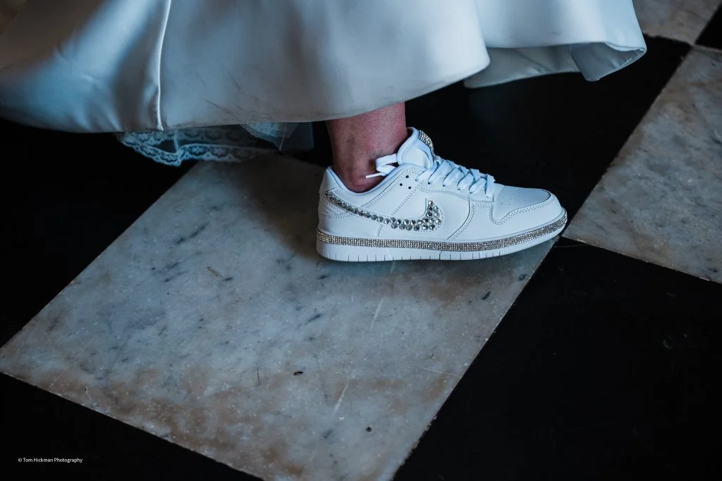 Close-up of a person wearing white sneakers with rhinestones and lace, partly visible, standing on a stone tile next to a white satin fabric.