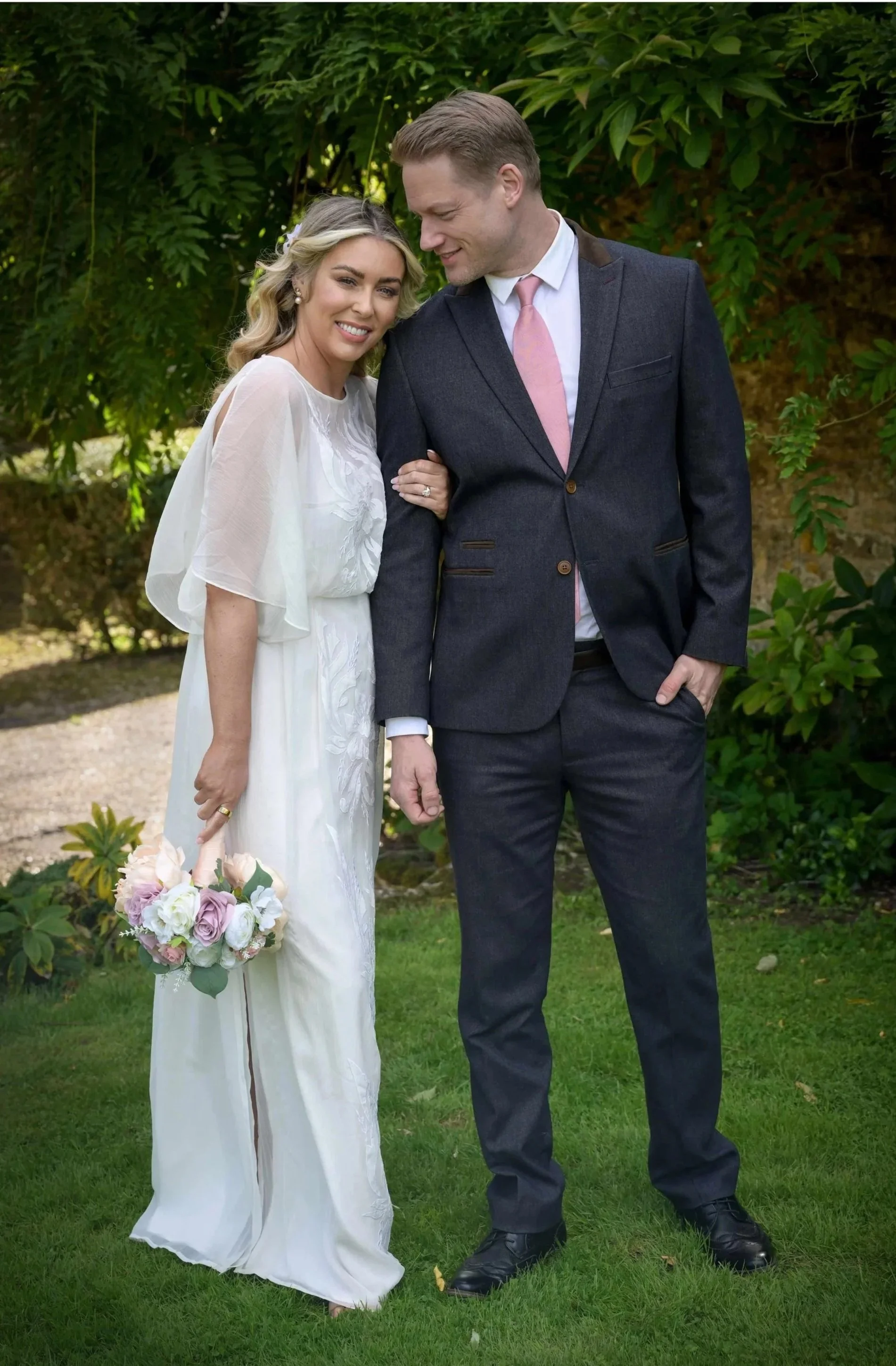 A bride in a white dress holding a bouquet of pink and white roses, smiling with a groom in a dark suit and pink tie, standing outdoors on green grass with trees in the background.