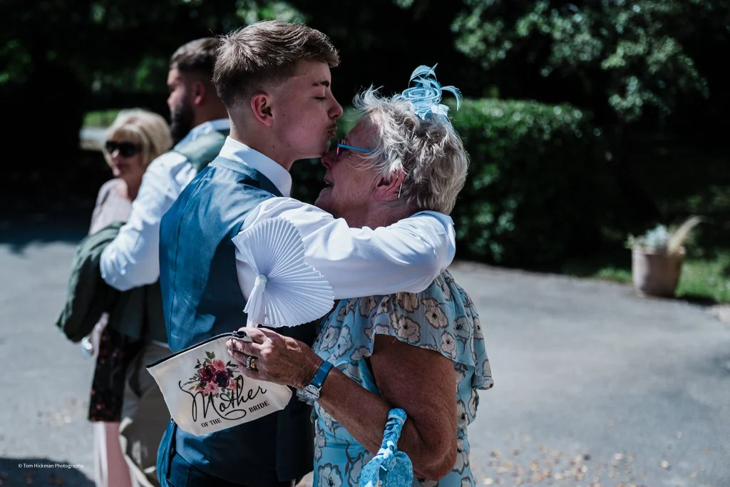 A young man in a blue vest and white shirt kisses an elderly woman on the forehead as they hug outdoors, with two people in the background, sunny weather, and greenery.