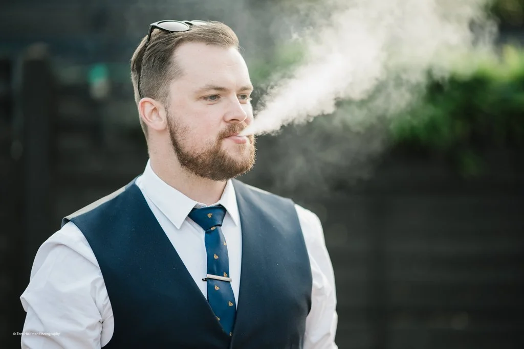 A man with a beard and glasses on his head blowing out smoke from his mouth outdoors during daytime, with greenery in the background.