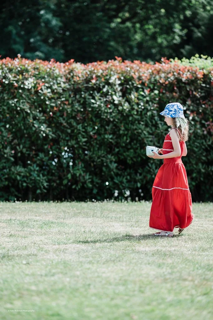A young girl in a red dress and a blue patterned sunhat walking on grass while holding a small white device, with a hedge and trees in the background.