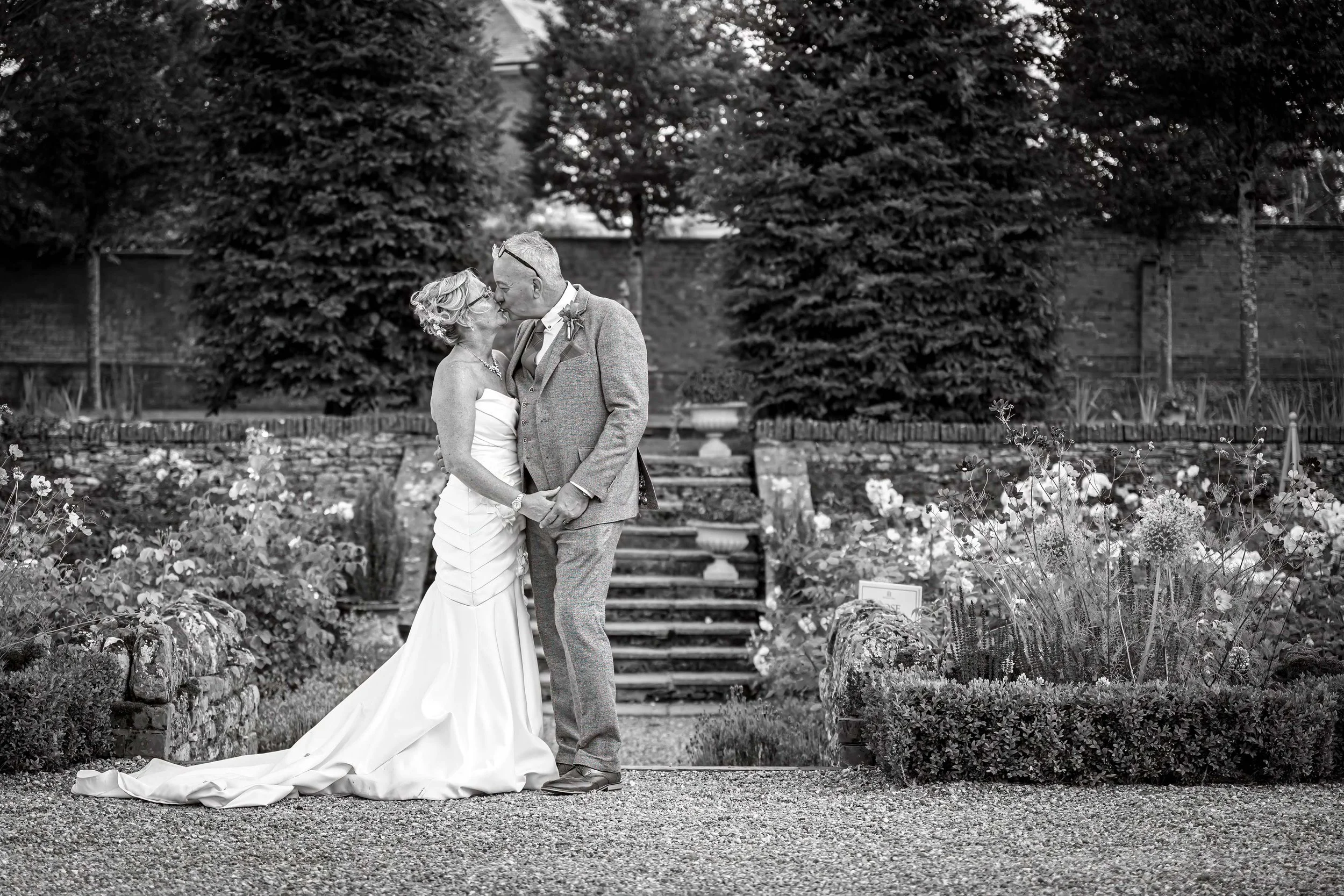 A bride and groom sharing a kiss in a garden setting during their wedding, with trees and a brick wall in the background.