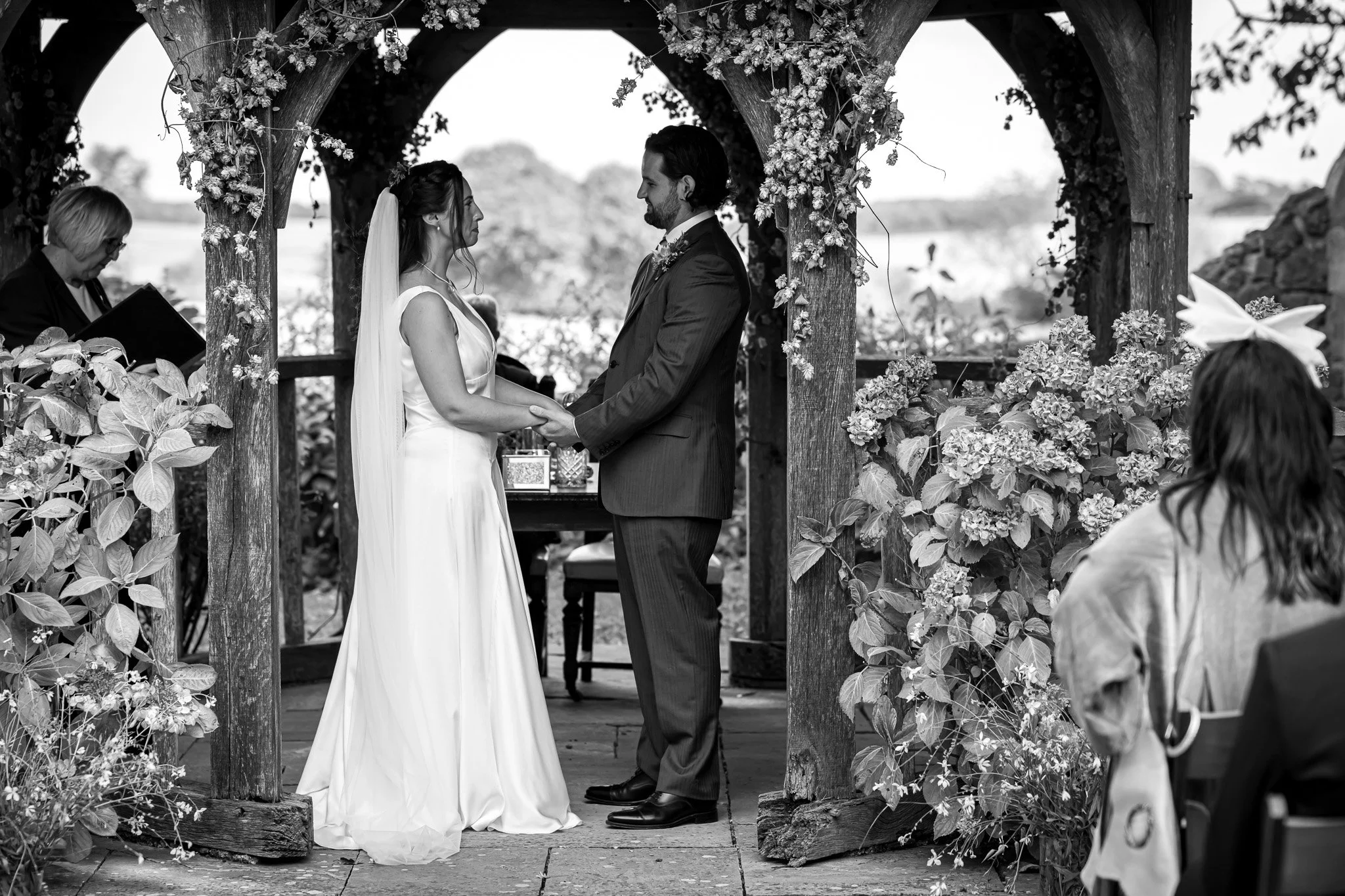 A black-and-white photo of a wedding ceremony at Solton Manor with a bride and groom holding hands and facing each other under a wooden arch decorated with flowers. An officiant stands nearby, and guests are visible in the foreground and background.