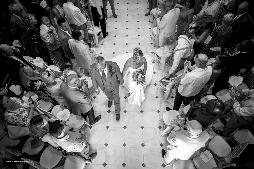 Black and white photo of a wedding ceremony with the bride and groom walking down the aisle, surrounded by seated and standing guests.