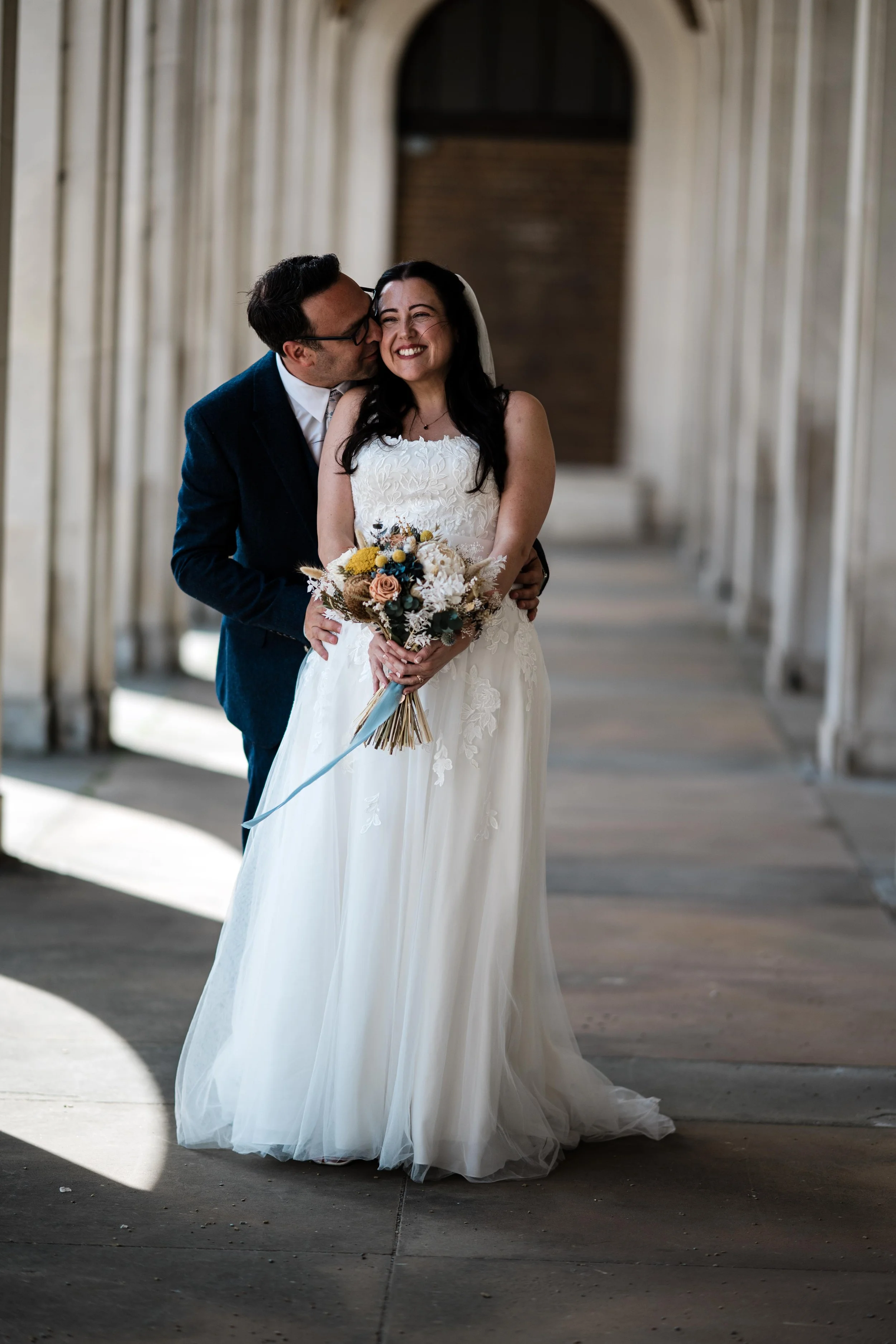 A bride and groom in wedding attire sharing a joyful moment, with the groom kissing the bride's cheek and the bride smiling, holding a bouquet of flowers, all inside a corridor with arched openings and hanging curtains.