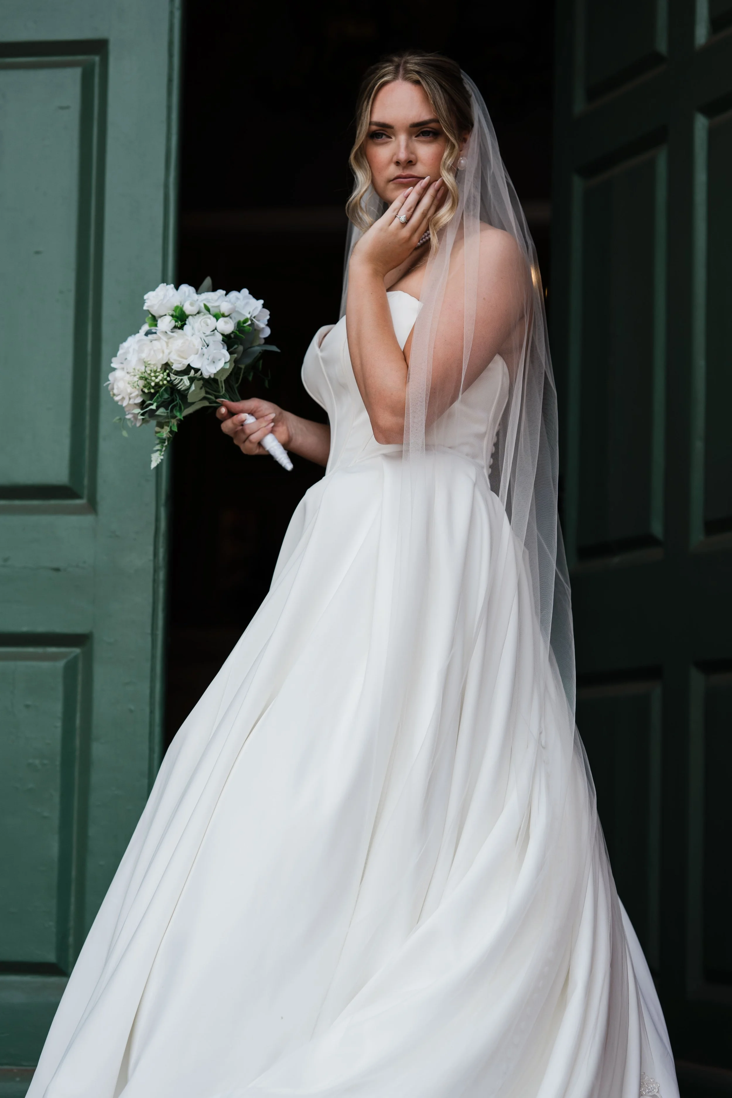 A woman in a wedding dress and veil holding a bouquet of white flowers, standing by green door, touching chin and looking to side.