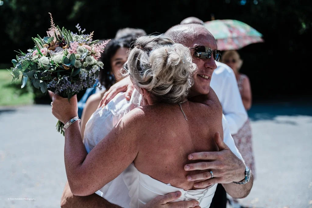 A happy elderly woman and a man are sharing an embrace, with the woman holding a large bouquet of flowers. They are outdoors, with other people in the background, one holding an umbrella.